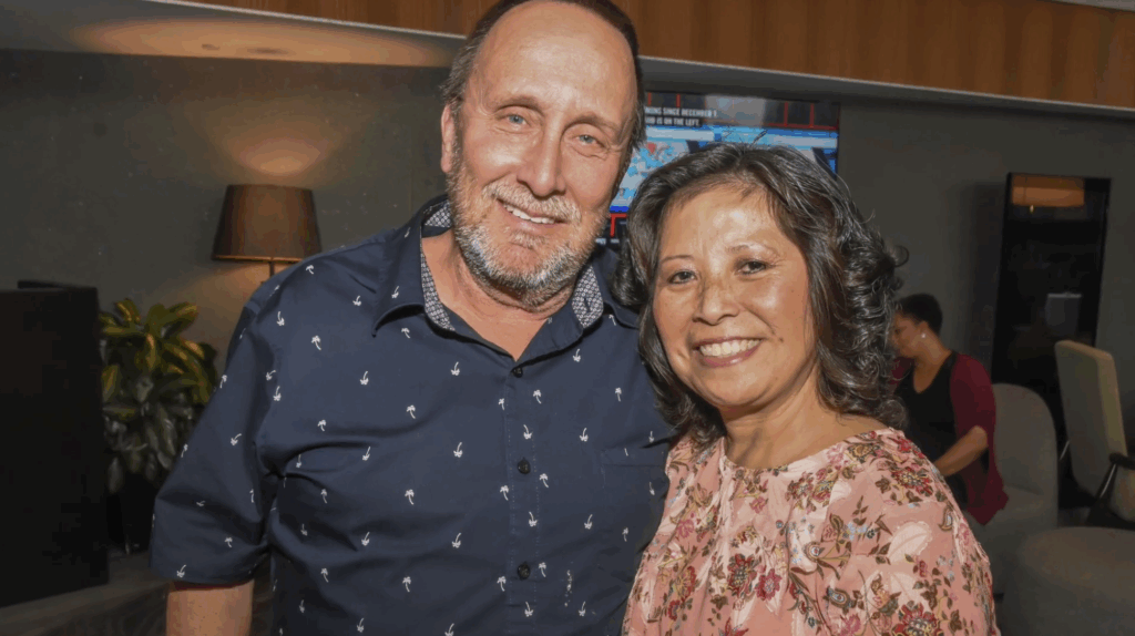 A man in a navy patterned shirt and a woman in a pink floral blouse smile together indoors, with other people and furniture in the background. | The DONNA Foundation