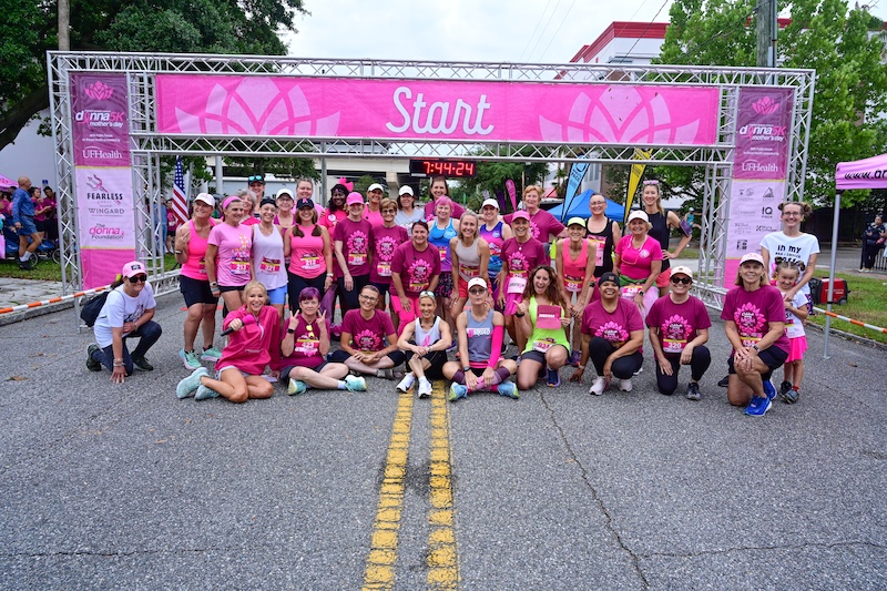 A group of runners in pink athletic wear pose together at a race starting line under a pink "Start" banner on a street. | The DONNA Foundation