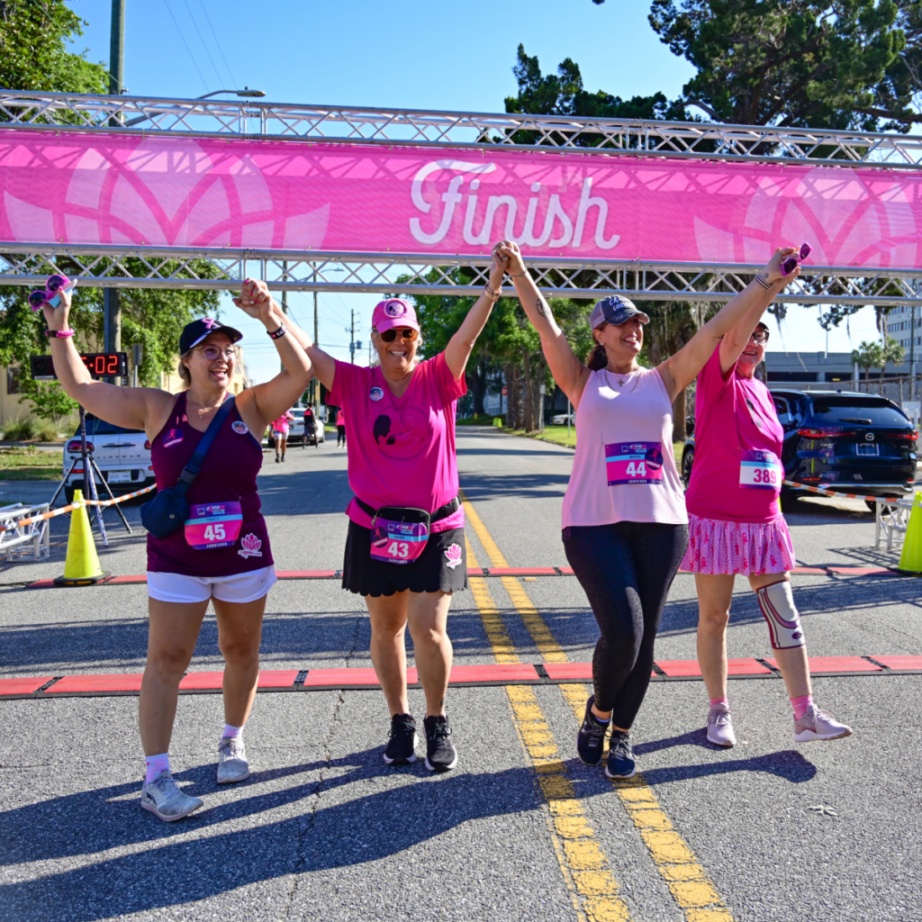Four women in athletic wear cross a pink "Finish" line outdoors, holding hands and celebrating after a race event. | The DONNA Foundation