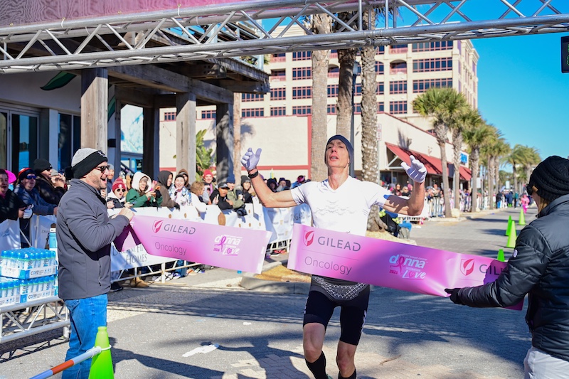A runner crosses the finish line of a race, raising his arms in triumph. Spectators watch from behind barriers, and event staff hold a pink finish line banner. | The DONNA Foundation