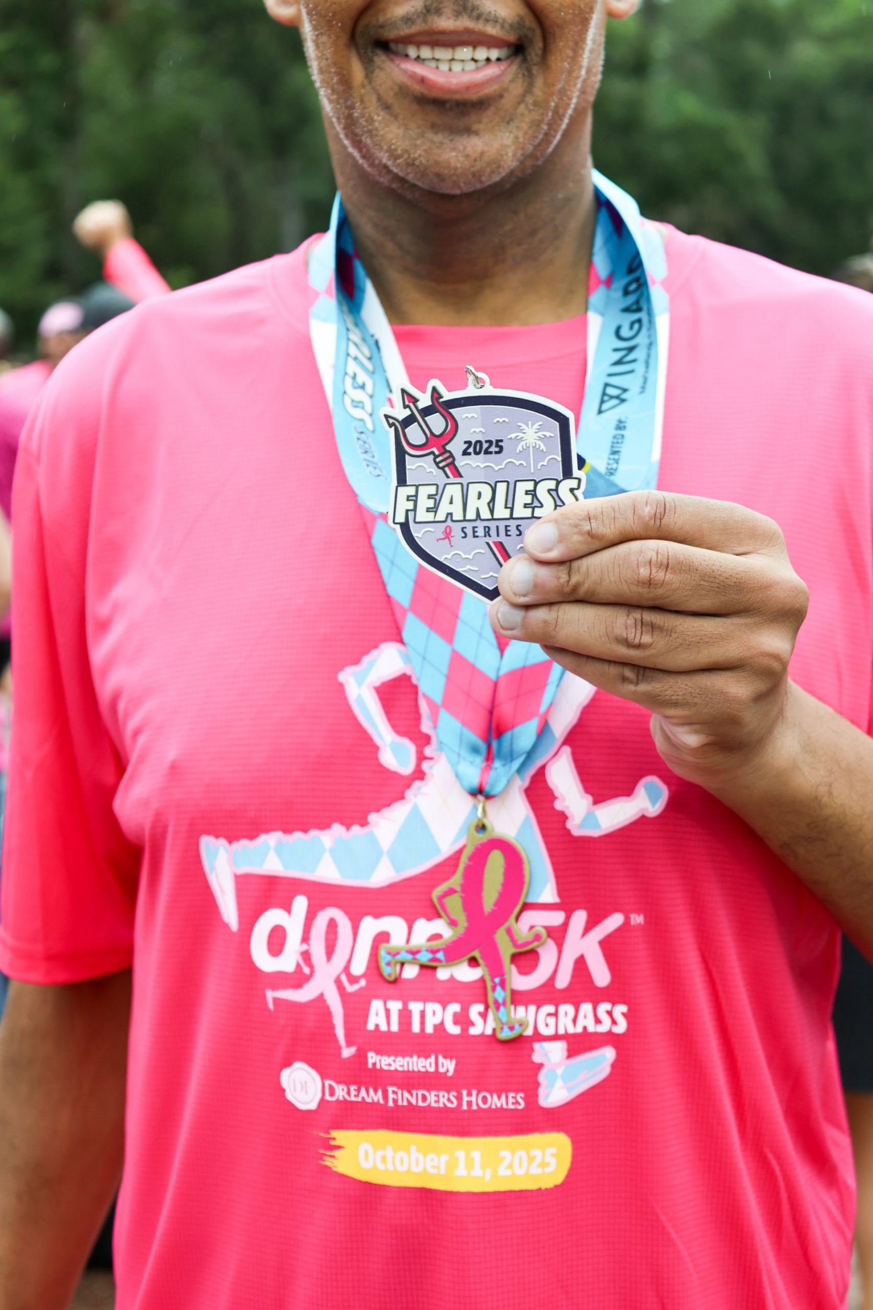 A person in a pink shirt holds up a "Fearless Series 2025" medal from a 5K event at TPC Sawgrass, with the race date of October 11, 2025, visible on the shirt. | The DONNA Foundation